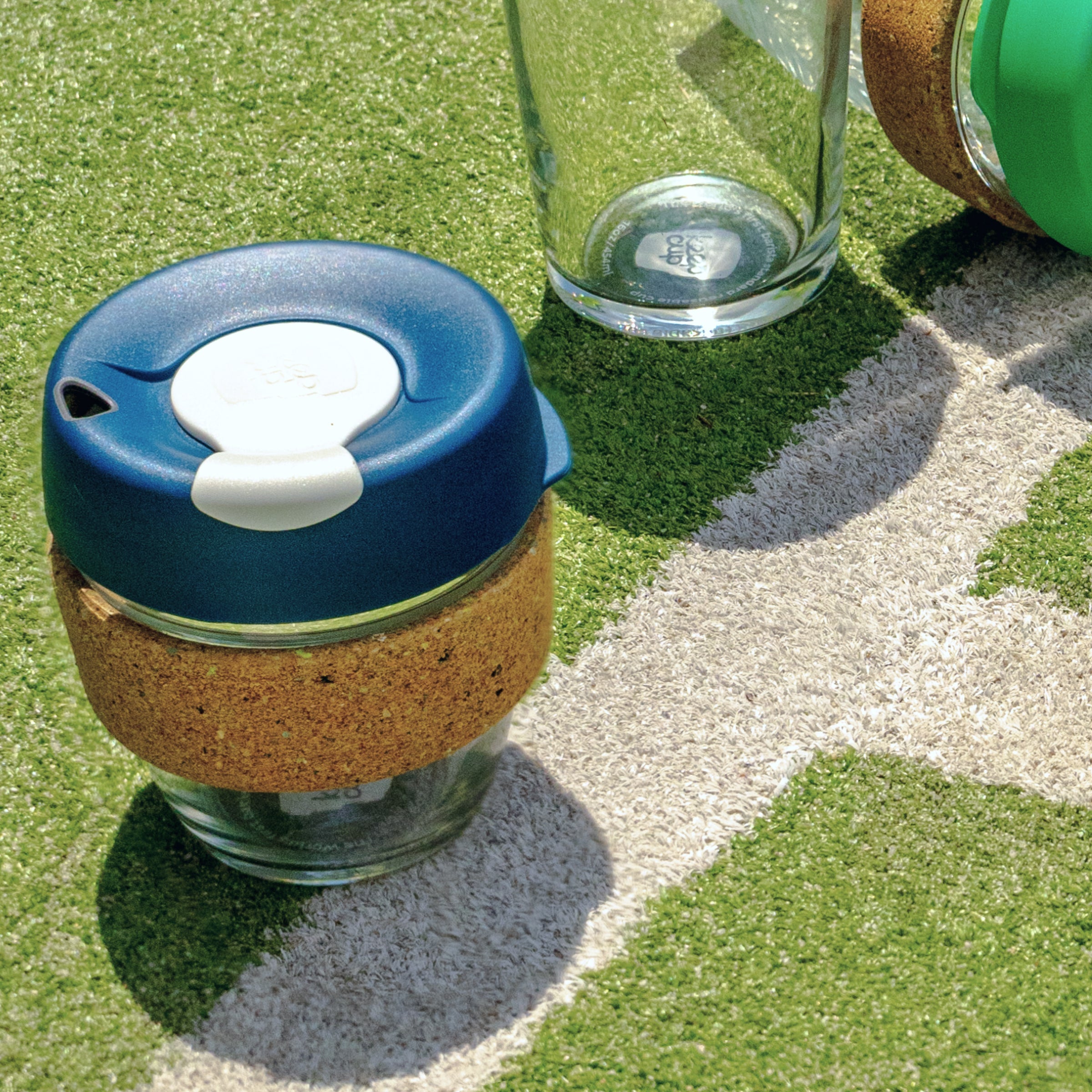 KeepCup Brew Cork Ace in S 8oz Rally, featuring a navy blue lid with white plug and natural cork band, placed on artificial grass turf with white pitch markings, alongside other glass KeepCup products in bright sunlight.