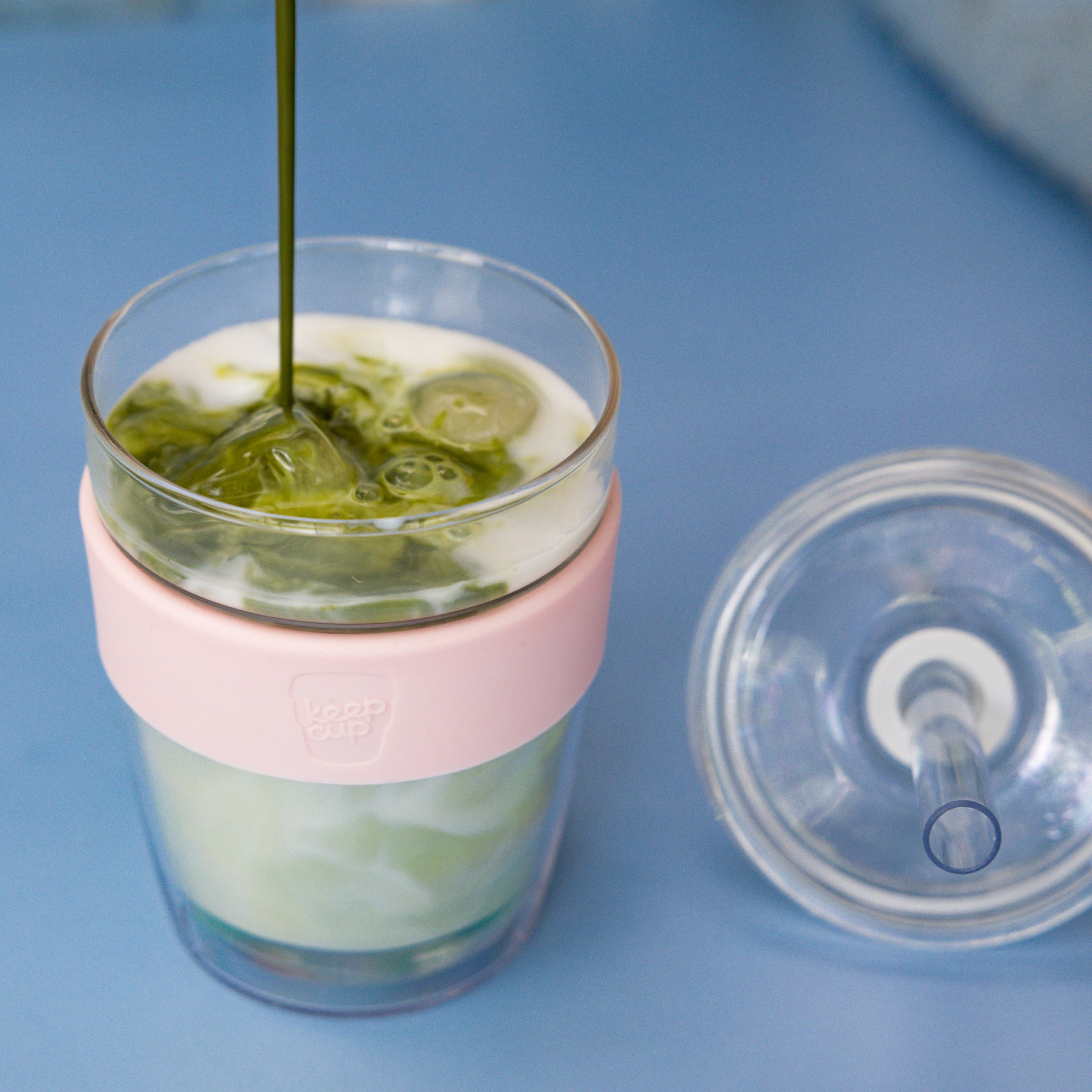 Glass mug with pink sleeve and clear lid on a blue background, containing a iced matcha.