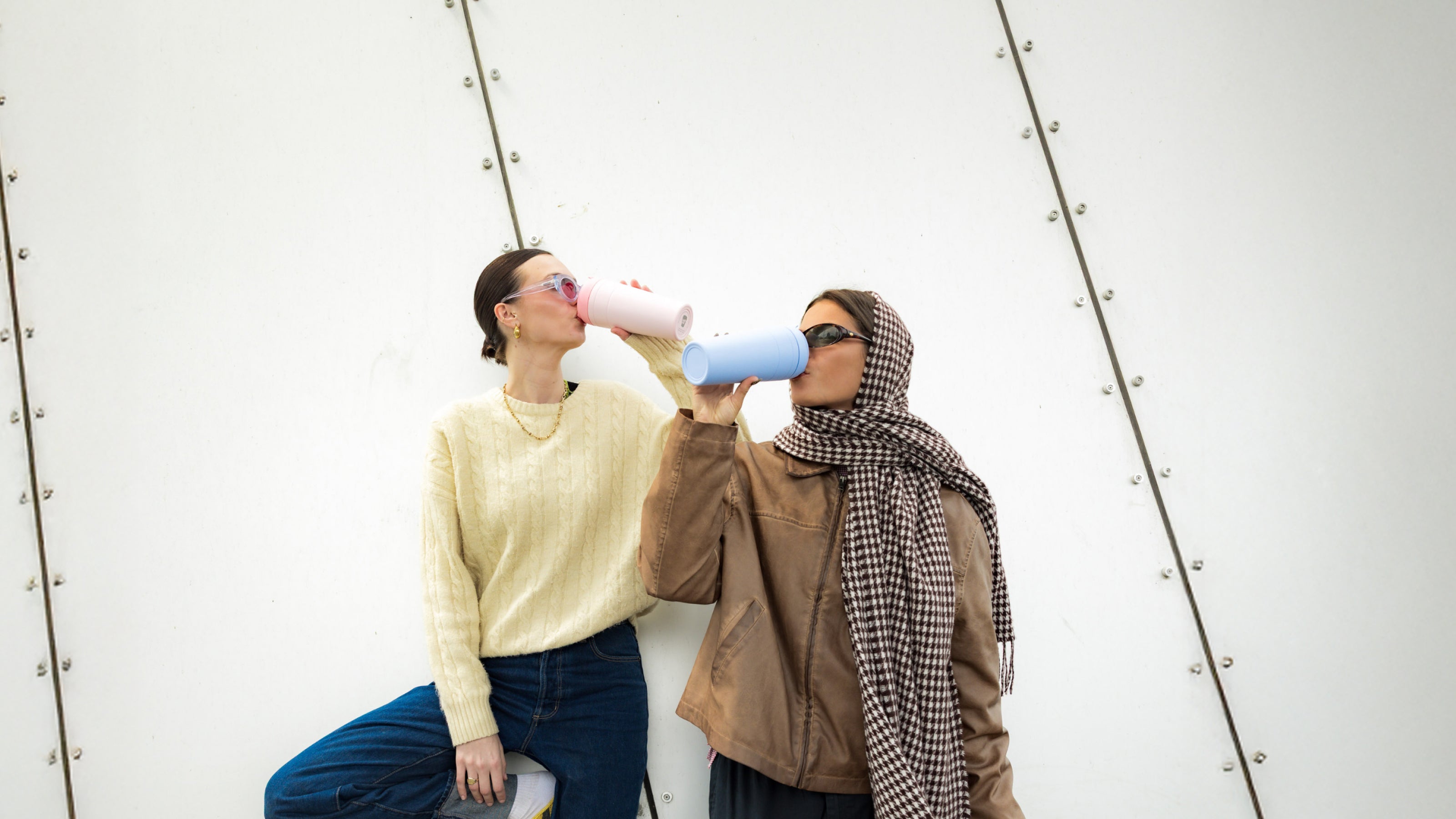 Two women drinking from reusable water bottles - one white, one blue - against white metal wall background
