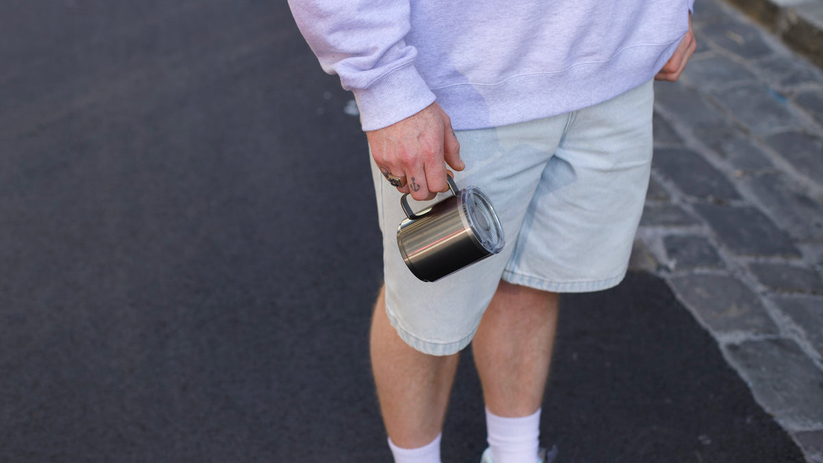 Person holding stainless steel reusable coffee cup with clear lid, wearing casual grey clothing and white socks