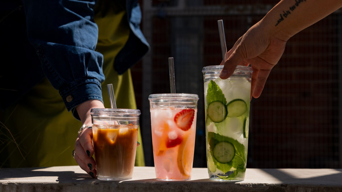Three glass tumblers with metal straws - iced coffee, strawberry drink, and cucumber mint water being held by hands