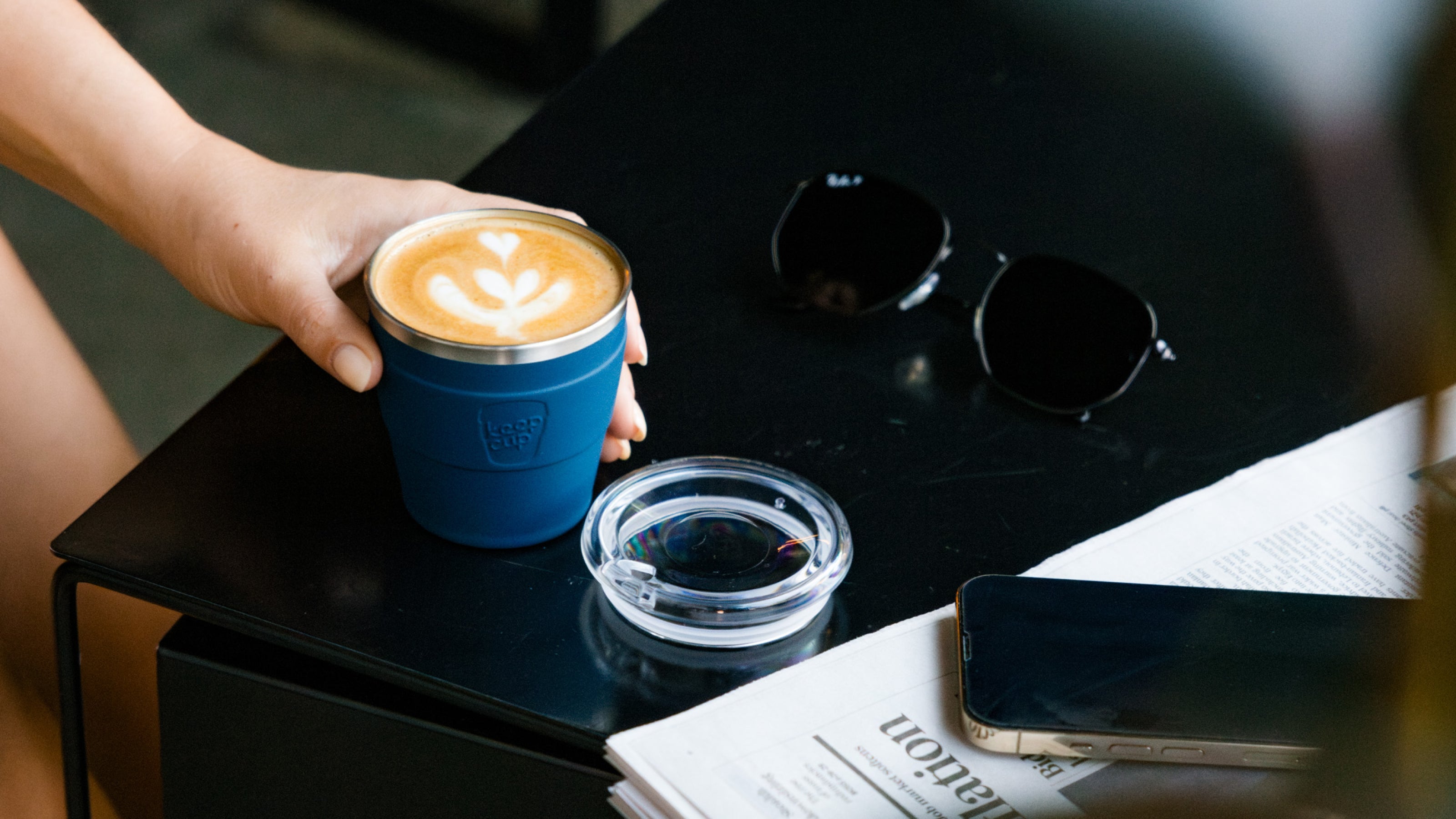 Person holding blue reusable coffee cup with latte art on black table with newspapers and sunglasses