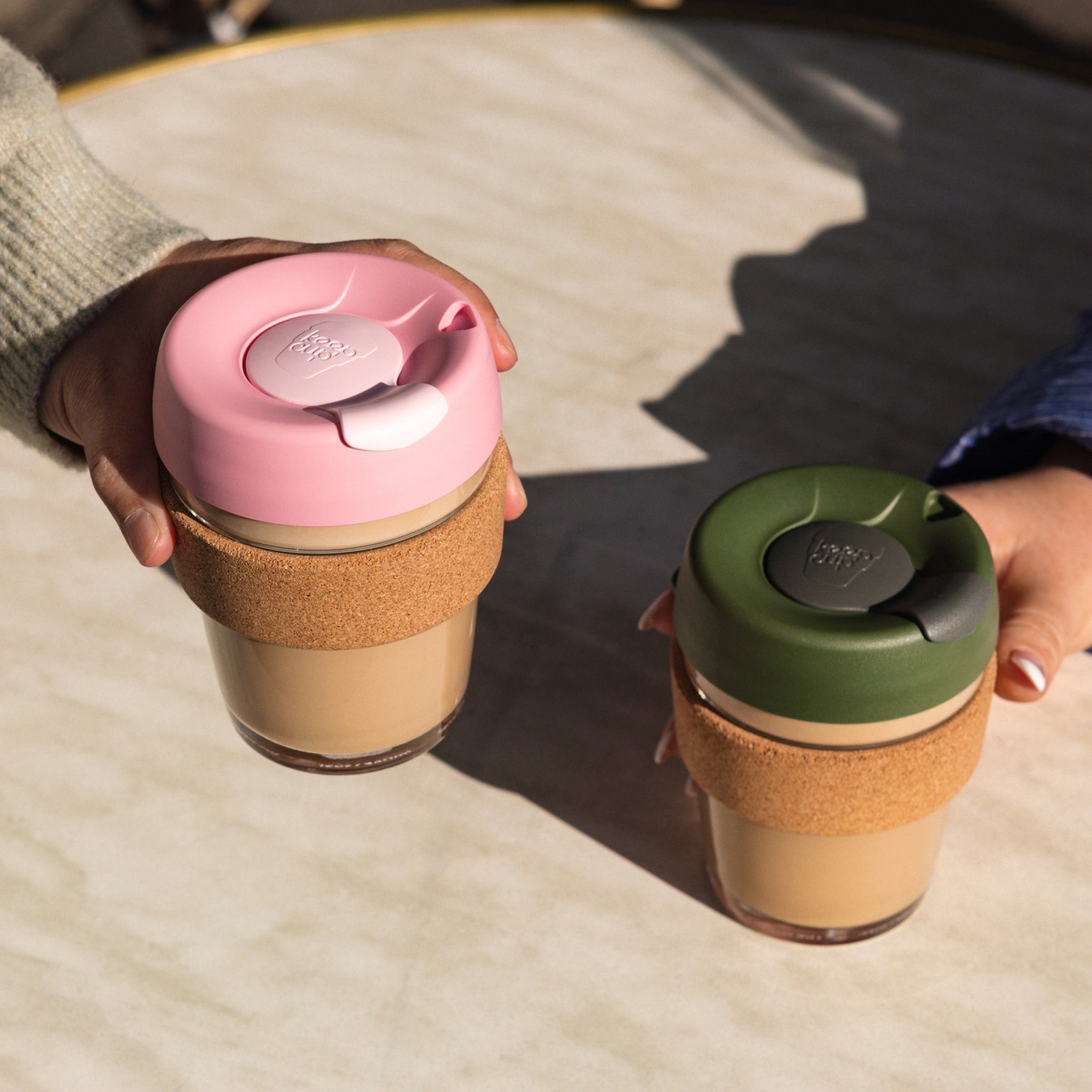 Two Keepcup reusable coffee cups with colorful lids held by hands on a wooden surface.