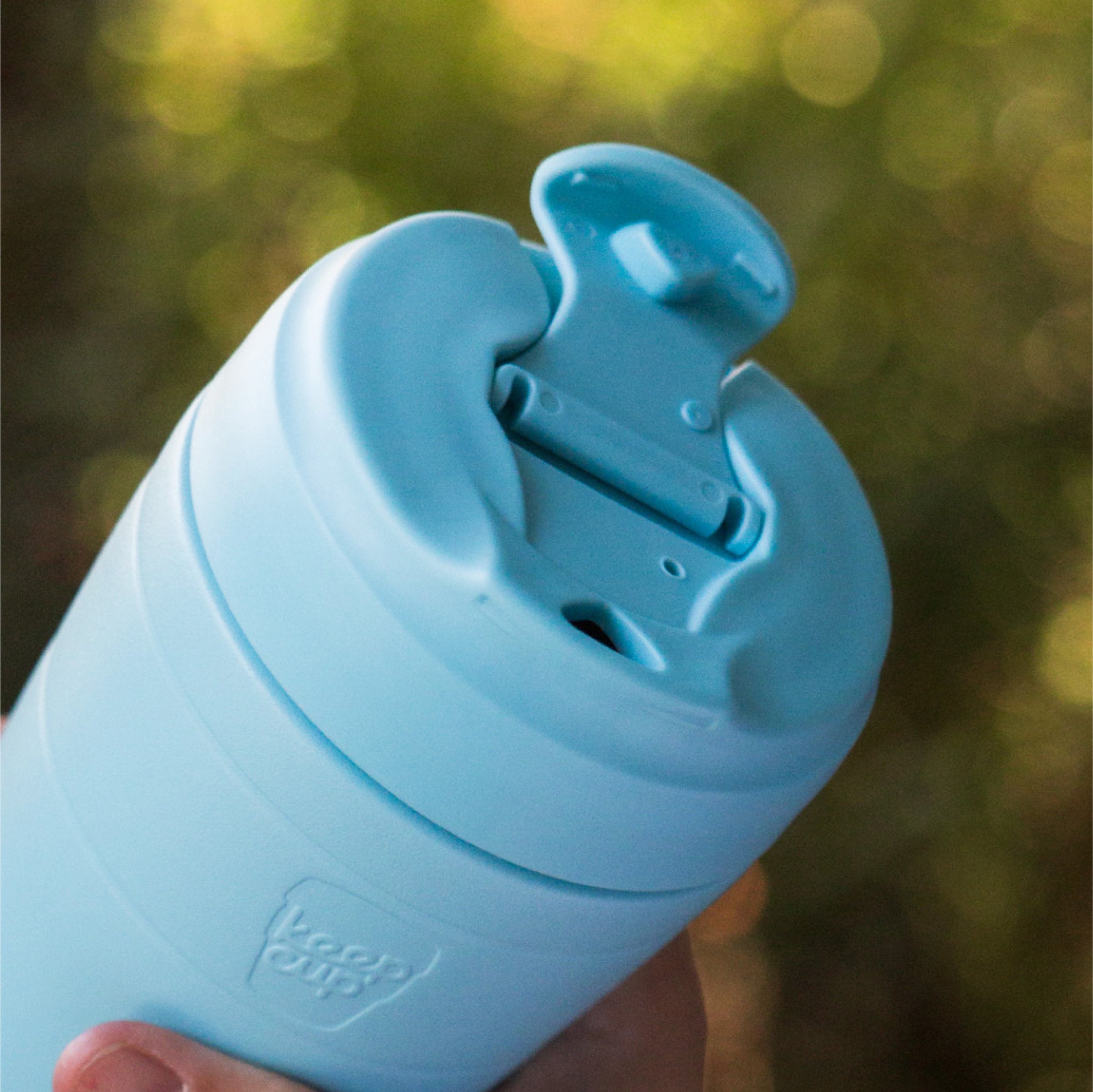 Close-up of a KeepCup Commuter travel mug with open sipper lid against a blurred natural background