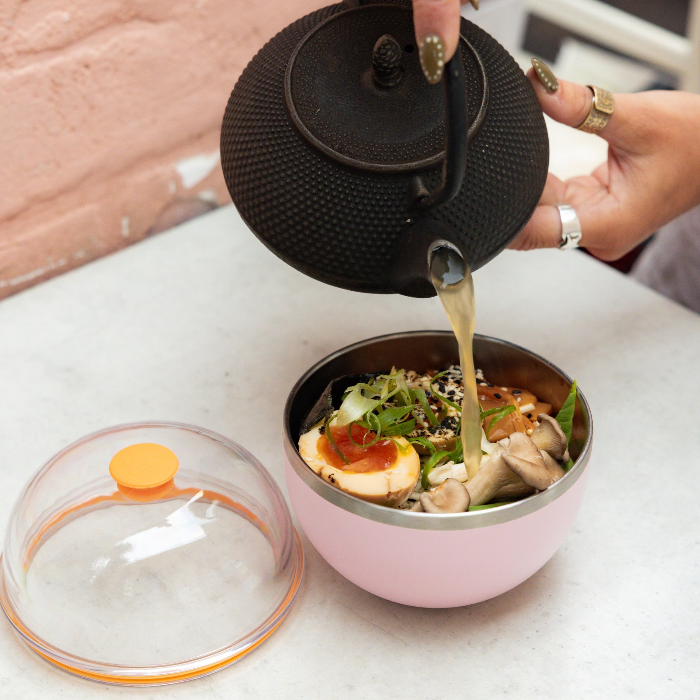 A hand pours broth from a black cast iron teapot into a KeepCup Go Bowl Luxe in Pashmak Pink, filled with a ramen-style dish featuring a soft-boiled egg, mushrooms, greens and sesame seeds. The clear Dome Lid with orange accent sits beside the bowl on a marble surface.