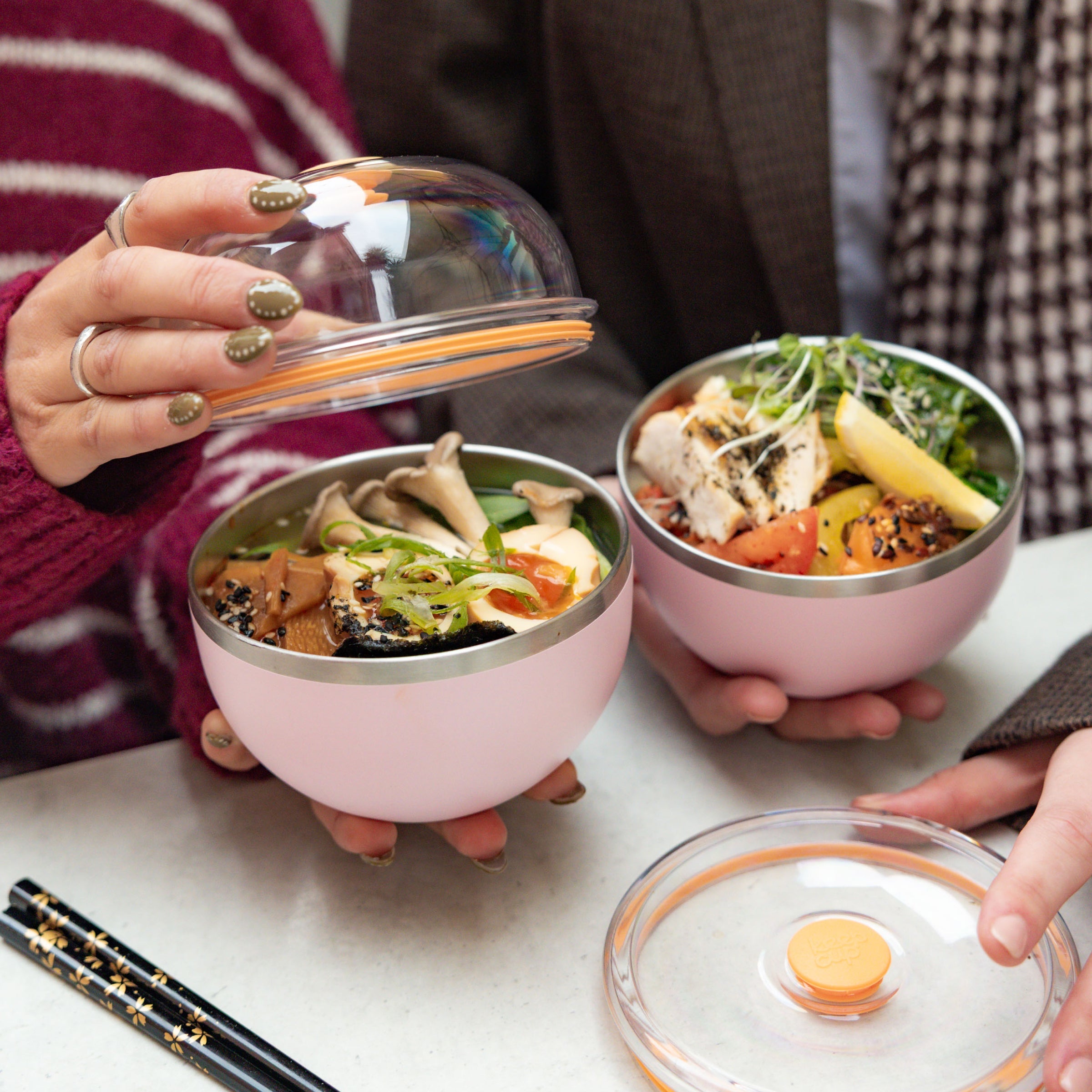 Two people sitting together opening their Go Bowl Luxe containers in Pashmak Pink, revealing colourful meals with mushrooms, tofu, chicken and fresh vegetables inside the stainless steel bowls, with dome lids and black chopsticks on the table.