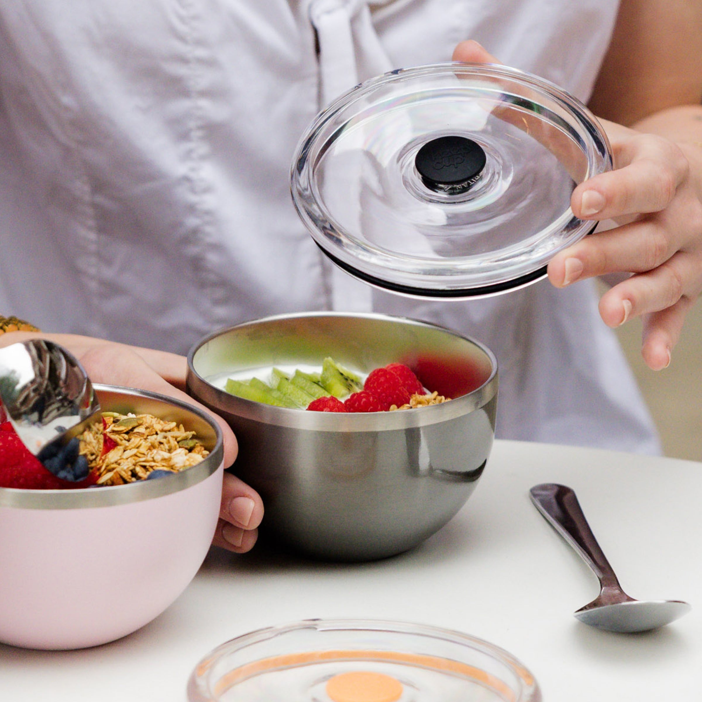 A person lifts the clear flat lid off the KeepCup Go Bowl Luxe in Nitro, revealing yoghurt topped with kiwi, raspberries and granola inside the stainless steel bowl, while holding a pink Go Bowl in their other hand, with a spoon resting on the white table.