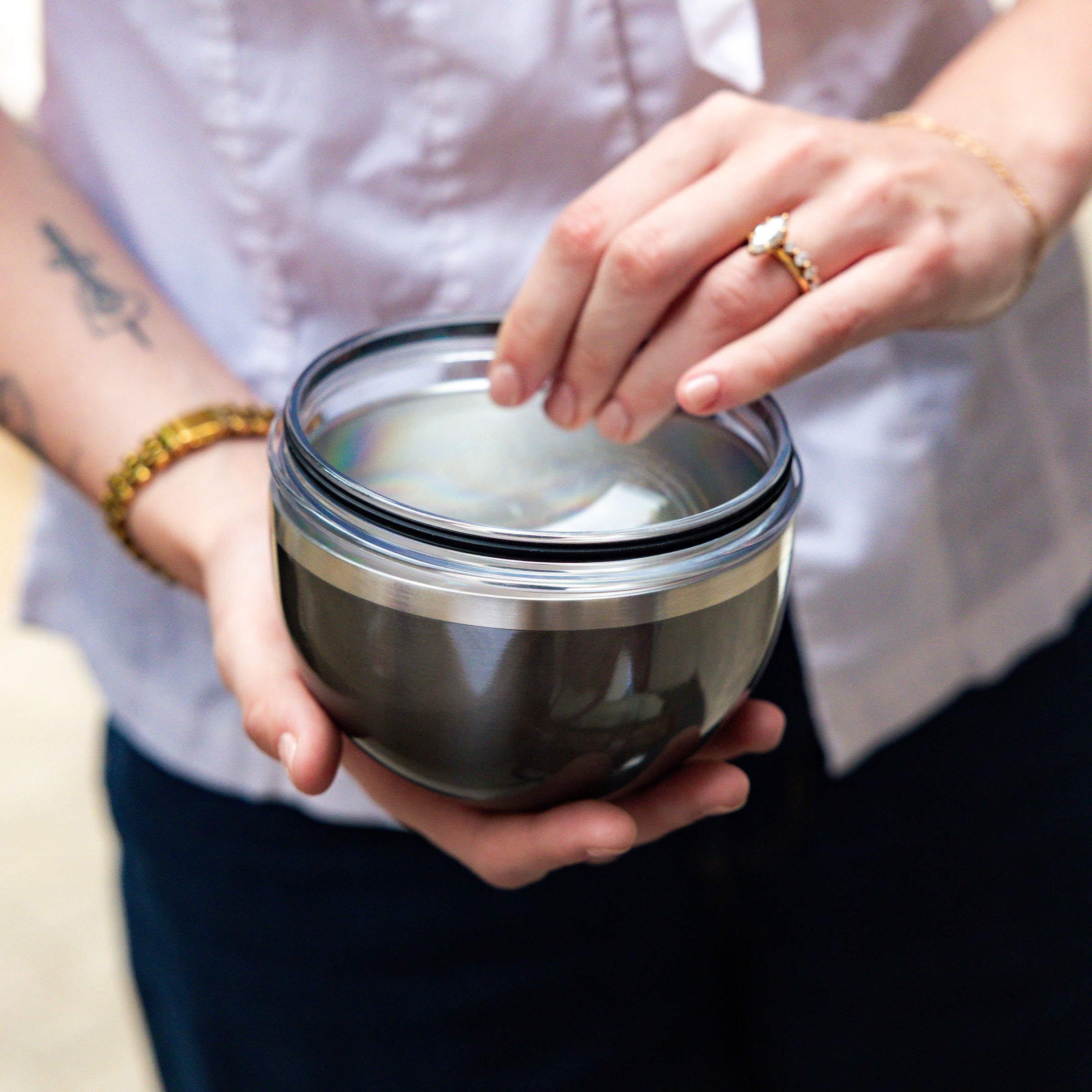 Hands holding a KeepCup Go Bowl Luxe in Nitro, with fingers lifting the clear dome lid to reveal the stainless steel bowl beneath, worn with gold jewellery and a light purple shirt.