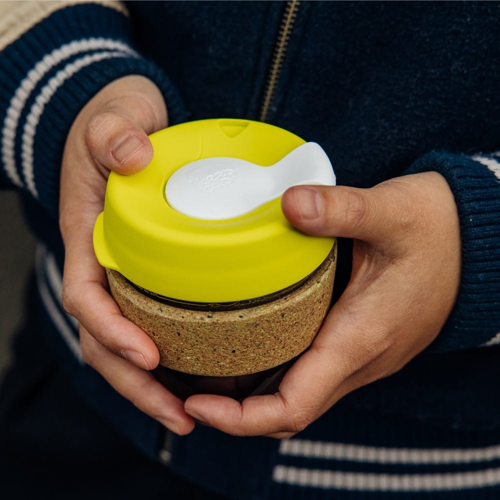 Person holding yellow and white reusable coffee cup with cork sleeve in both hands, wearing navy striped jumper