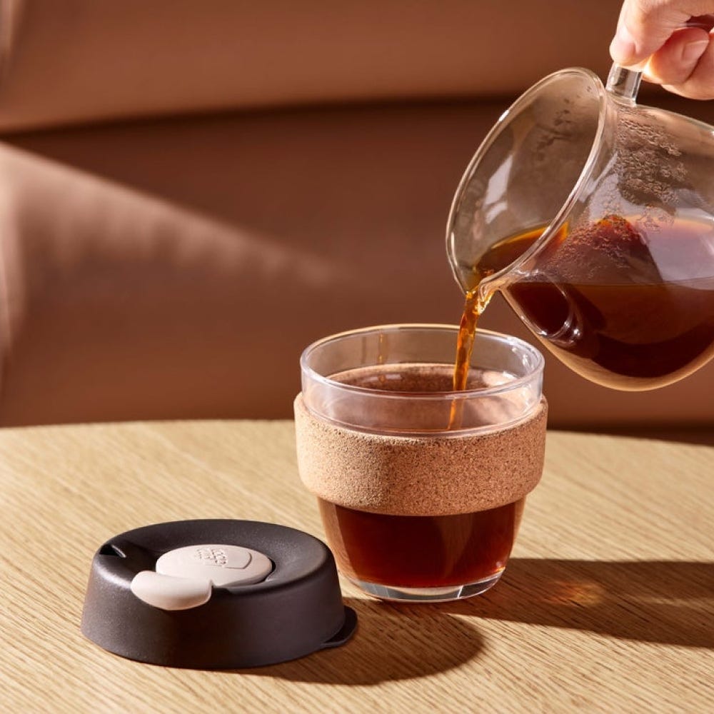 Pouring coffee from a glass pot into a glass with cork band on a wooden surface.