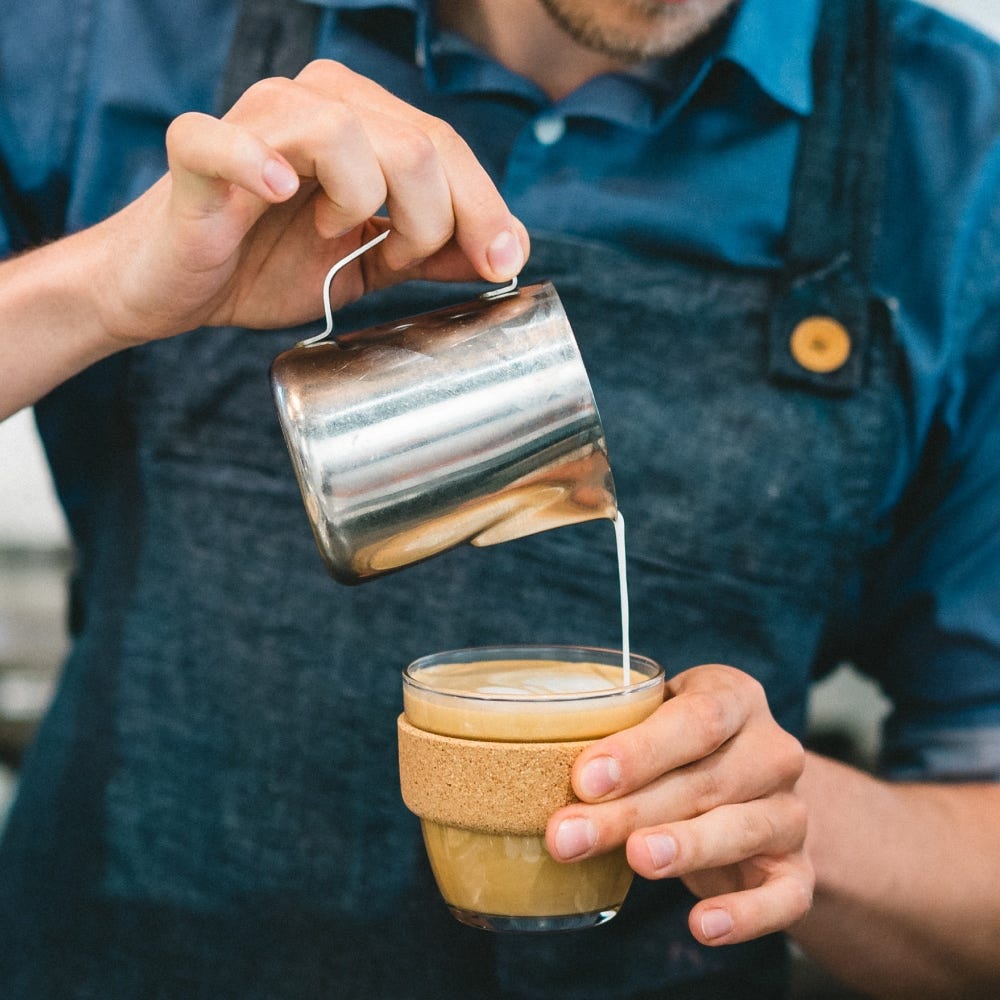 Person pouring milk into a glass of coffee with cork band using a stainless steel pitcher.
