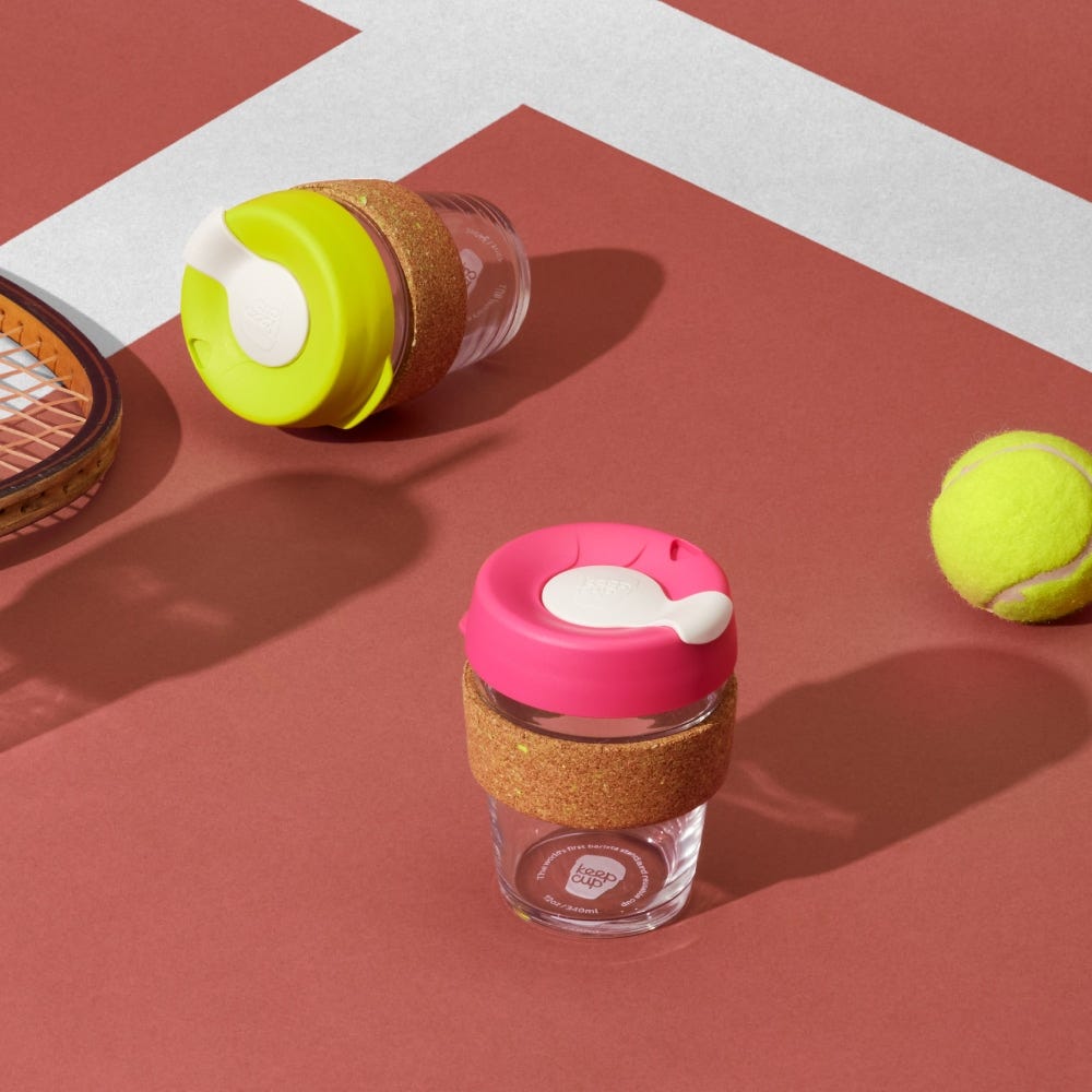 Two KeepCup reusable coffee cups with cork bands - one bright green lid, one pink lid - displayed with tennis racquet and ball on red court surface