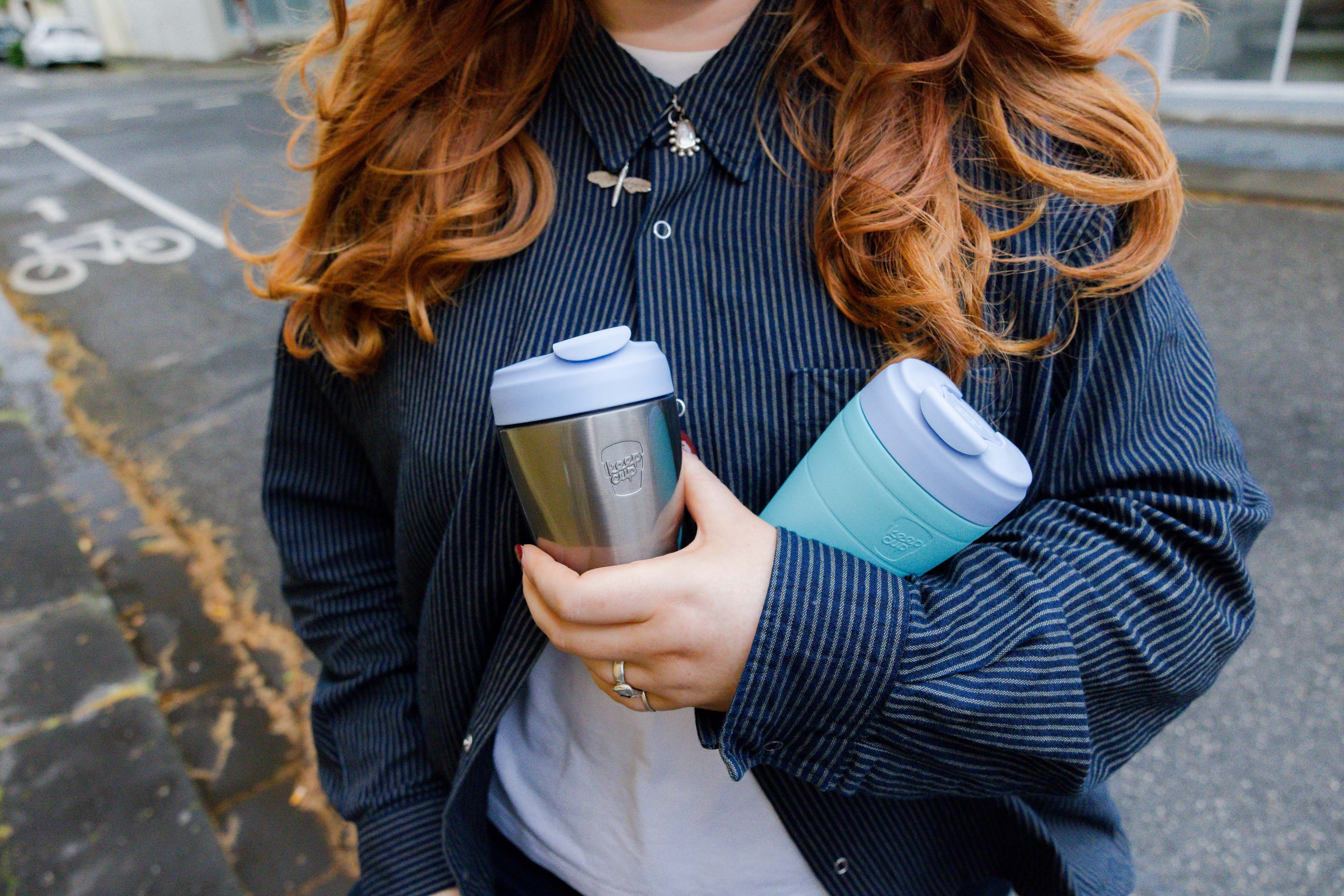 Person holding two insulated travel mugs outdoors on a street.
