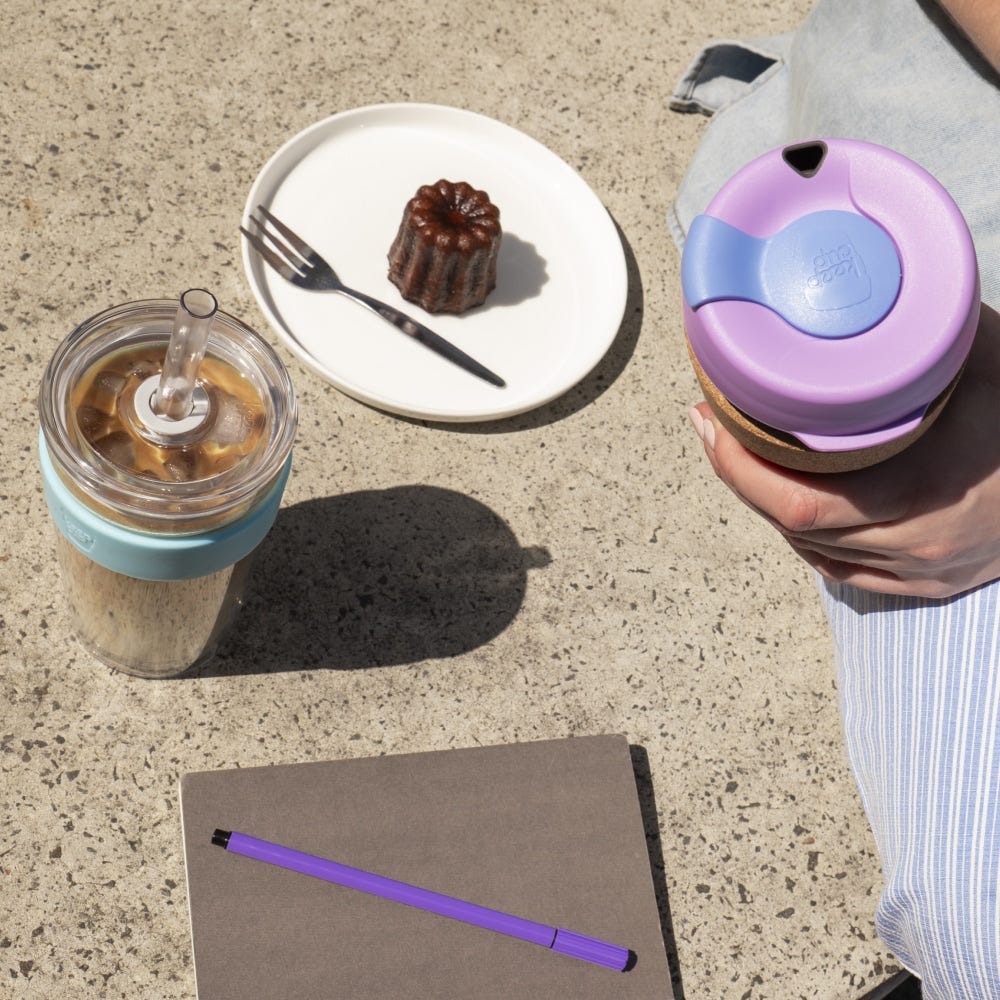 A hand holds the KeepCup Brew Cork XS 6oz in Moonlight, showing its lilac and maeve lid from above, beside an iced coffee in a teal KeepCup, a canelé on a white plate, and a grey notebook with purple pen on a sunlit concrete surface.
