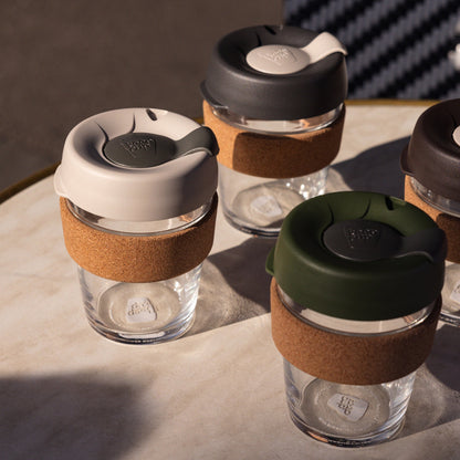 Four KeepCup Brew Cork reusable coffee cups in various colours displayed on a marble table in warm sunlight, featuring glass bodies with natural cork bands and lids, including the Filter colourway with its latte and soft charcoal lid