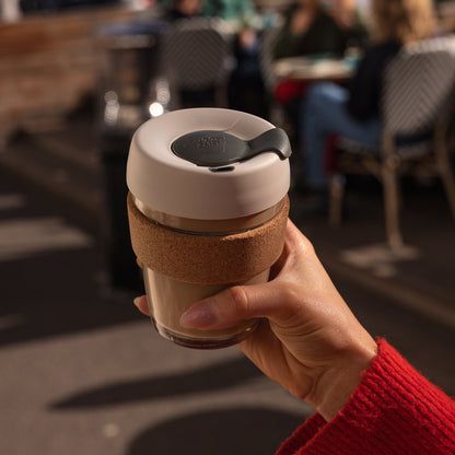 A hand in a red jumper holds a KeepCup Brew Cork M 12oz in Filter, showing the glass cup with cork band, latte coloured lid and soft charcoal sipper plug, with a blurred café setting in the background.