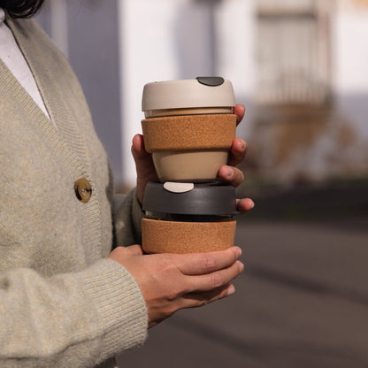 A person in a sage green cardigan holds two stacked KeepCup Brew Cork reusable coffee cups outdoors, featuring the S 8oz Filter colourway on top with a latte lid and cork band, and a darker variant beneath.
