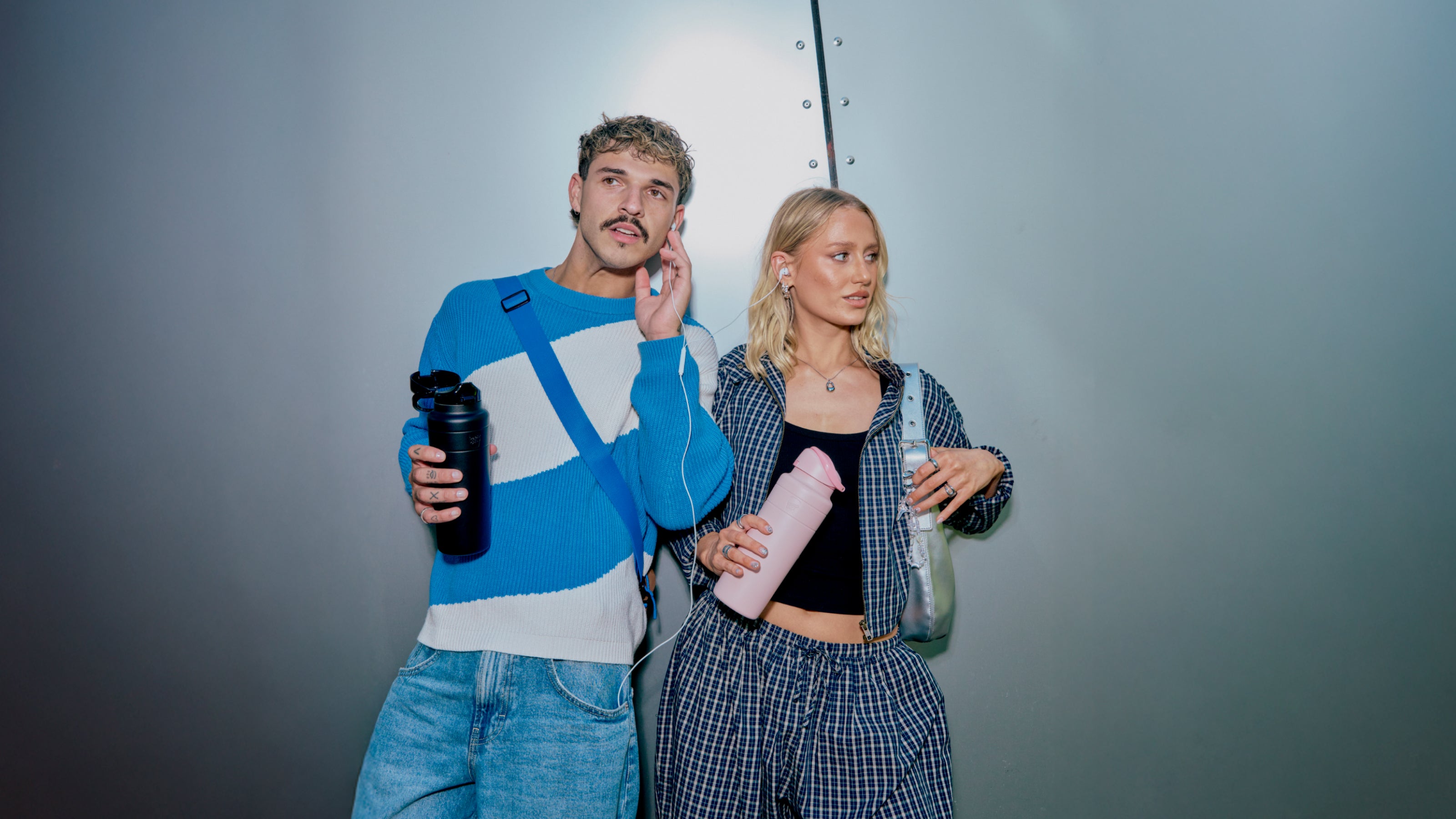 Two people standing side by side holding drinks against a plain background