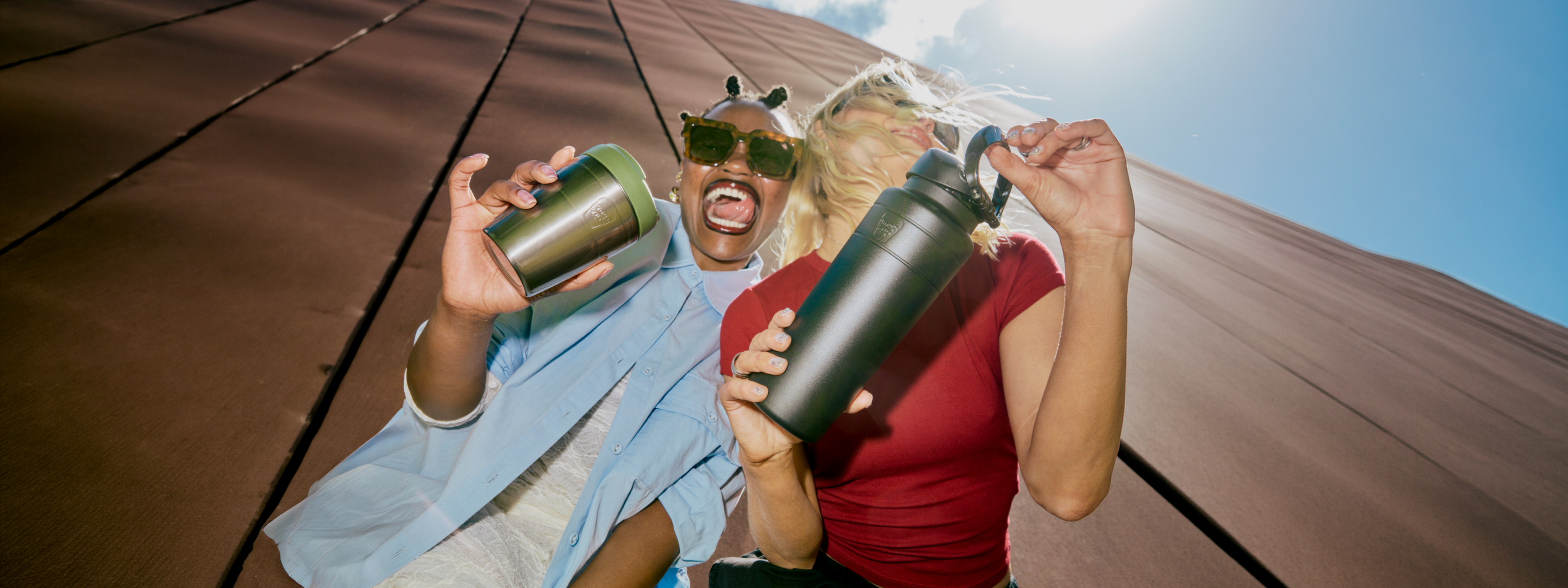 Two people outdoors on a sloped brown roof under a blue sky, one person holding a Commuter insulated travel cup and the other holding an Ora insulated water bottle.