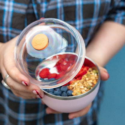 A person in a blue and grey plaid shirt holds an open KeepCup Go Bowl Luxe in Pashmak Pink, lifting the clear dome lid to reveal a healthy breakfast of blueberries, strawberries, yoghurt and granola inside the stainless steel bowl.