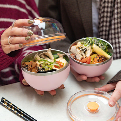 Two people sitting together opening their Go Bowl Luxe containers in Pashmak Pink, revealing colourful meals with mushrooms, tofu, chicken and fresh vegetables inside the stainless steel bowls, with dome lids and black chopsticks on the table.
