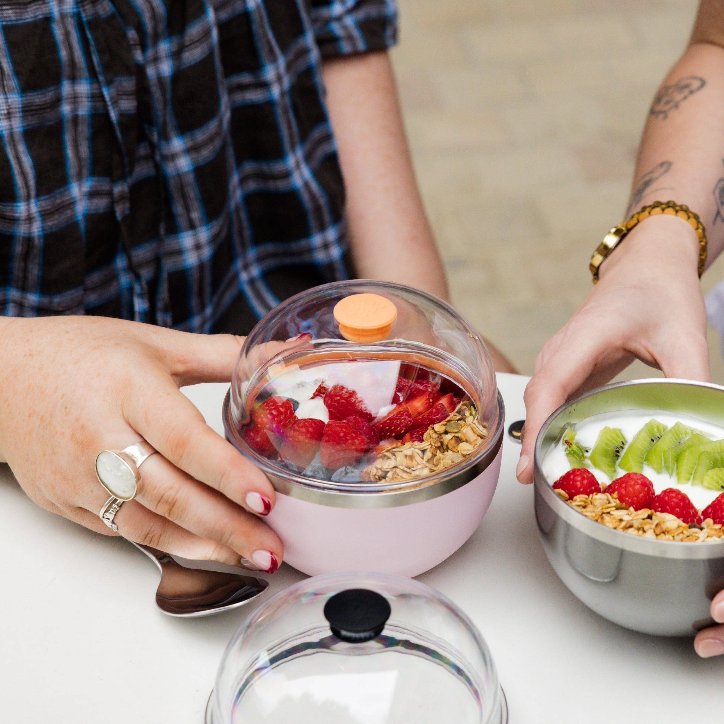 Two people sharing a meal, one holding a Go Bowl Luxe in Pashmak Pink with dome lid containing yoghurt, berries and granola, while the other holds an open stainless steel bowl with kiwi, strawberries and oats.