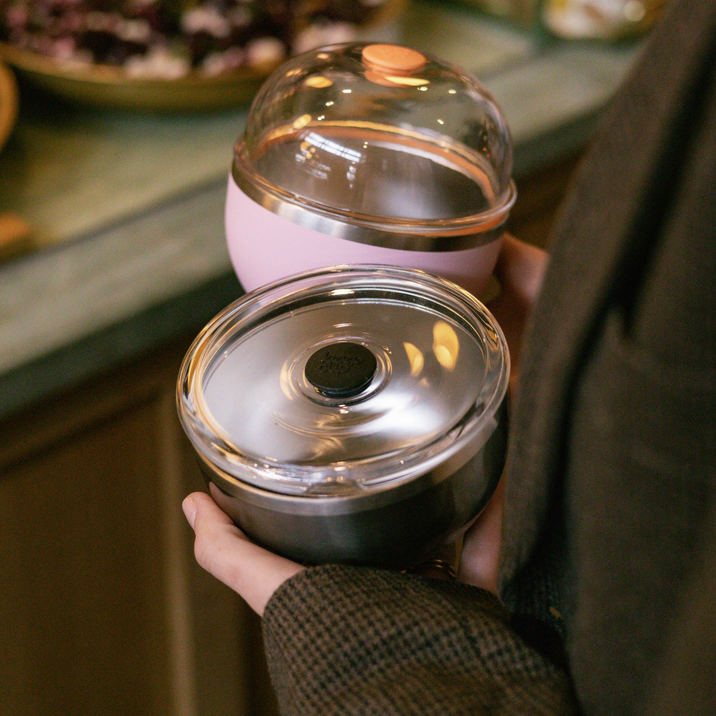 A hand holding the KeepCup Go Bowl Luxe in Nitro, featuring a dark base with stainless steel interior and clear flat lid, alongside a pink domed KeepCup, set against a café counter backdrop.