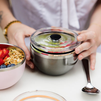 Hands placing the clear flat lid onto the KeepCup Go Bowl Luxe in Nitro, a stainless steel bowl containing granola and fresh fruit, with a pink bowl of berries and granola beside it on a white table.