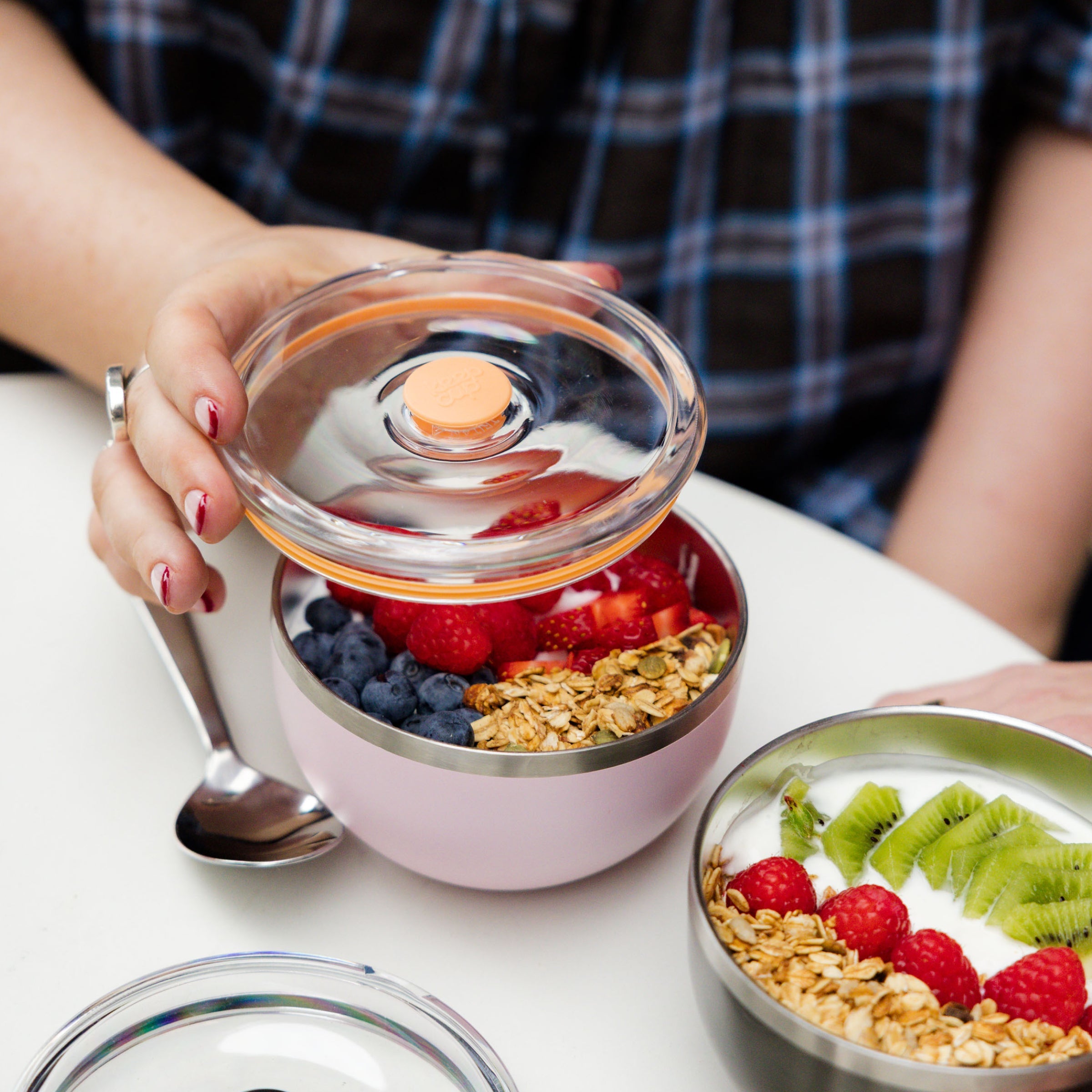 A hand with red nail polish lifts the clear flat lid off a KeepCup Go Bowl Luxe in Pashmak Pink, revealing layers of granola, blueberries, raspberries and strawberries inside, with a metal spoon resting against the bowl and another yoghurt bowl with kiwi and fruit visible on the white table.