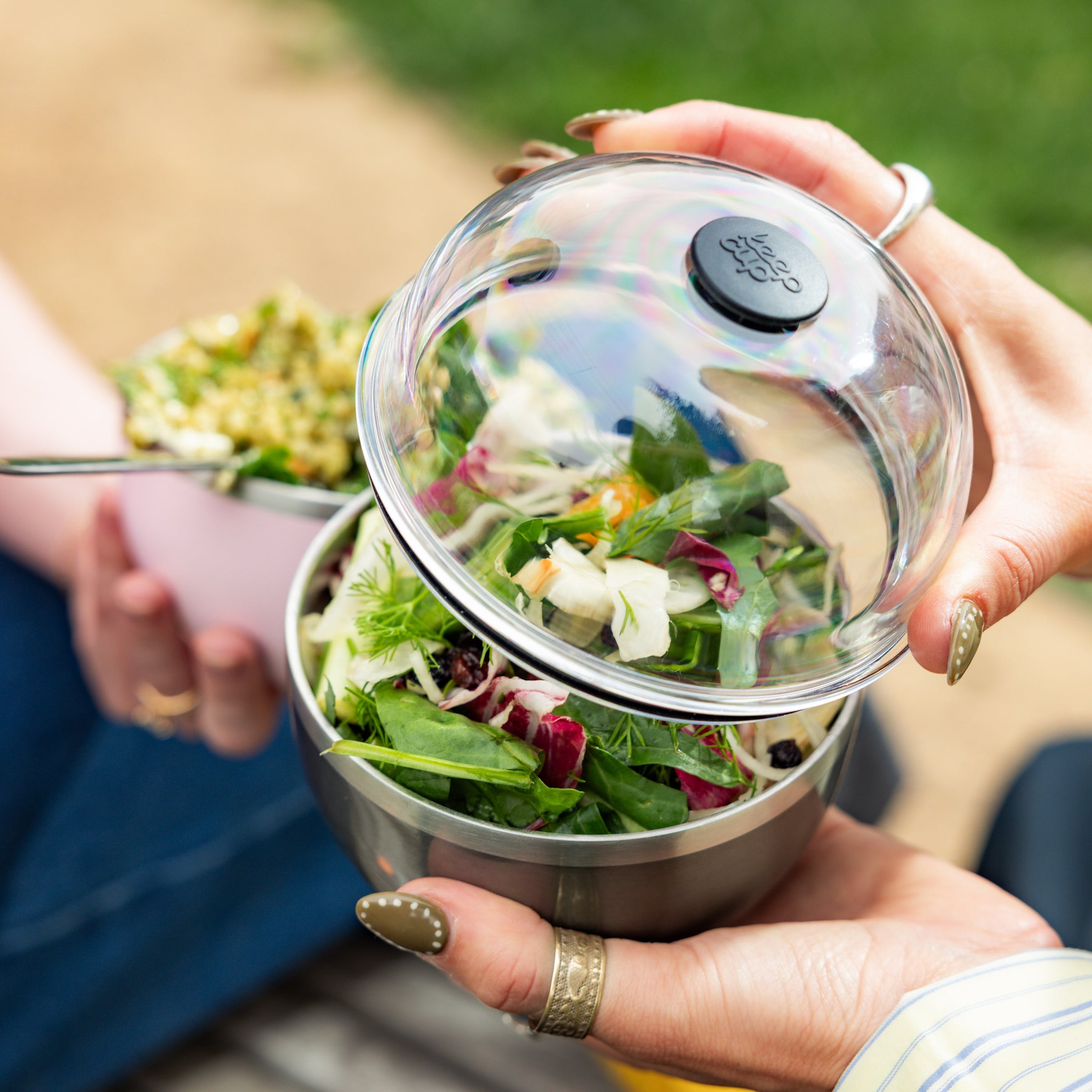 Hands holding an open KeepCup Go Bowl Luxe in Nitro, revealing a fresh mixed leaf salad inside the stainless steel bowl, with the clear dome lid lifted to show the colourful contents during an outdoor picnic setting.