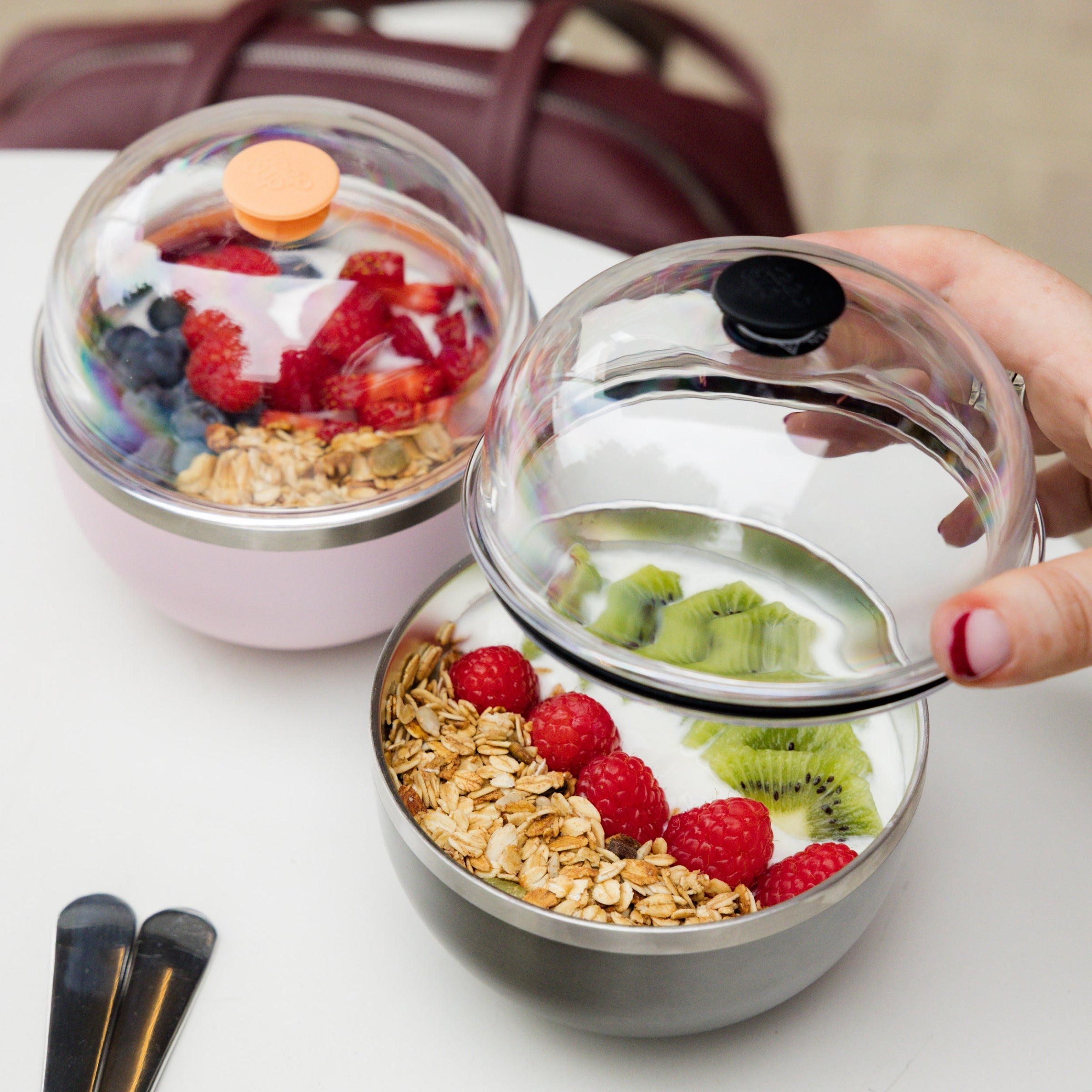 A hand with red nail polish lifts the clear dome lid off a Go Bowl Luxe in Nitro, revealing yoghurt topped with raspberries, kiwi slices and granola, while a second Go Bowl in pink with an orange lid sits in the background alongside a burgundy bag and black cutlery.