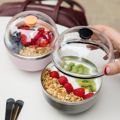 A hand with red nail polish lifts the clear dome lid off a Go Bowl Luxe in Nitro, revealing yoghurt topped with raspberries, kiwi slices and granola, while a second Go Bowl in pink with an orange lid sits in the background alongside a burgundy bag and black cutlery.