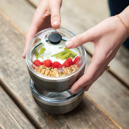 Hands holding a KeepCup Go Bowl Luxe in Nitro, featuring a clear dome lid and stainless steel band, filled with layered yoghurt, raspberries, kiwi and granola, stacked on a second bowl on a wooden outdoor table.