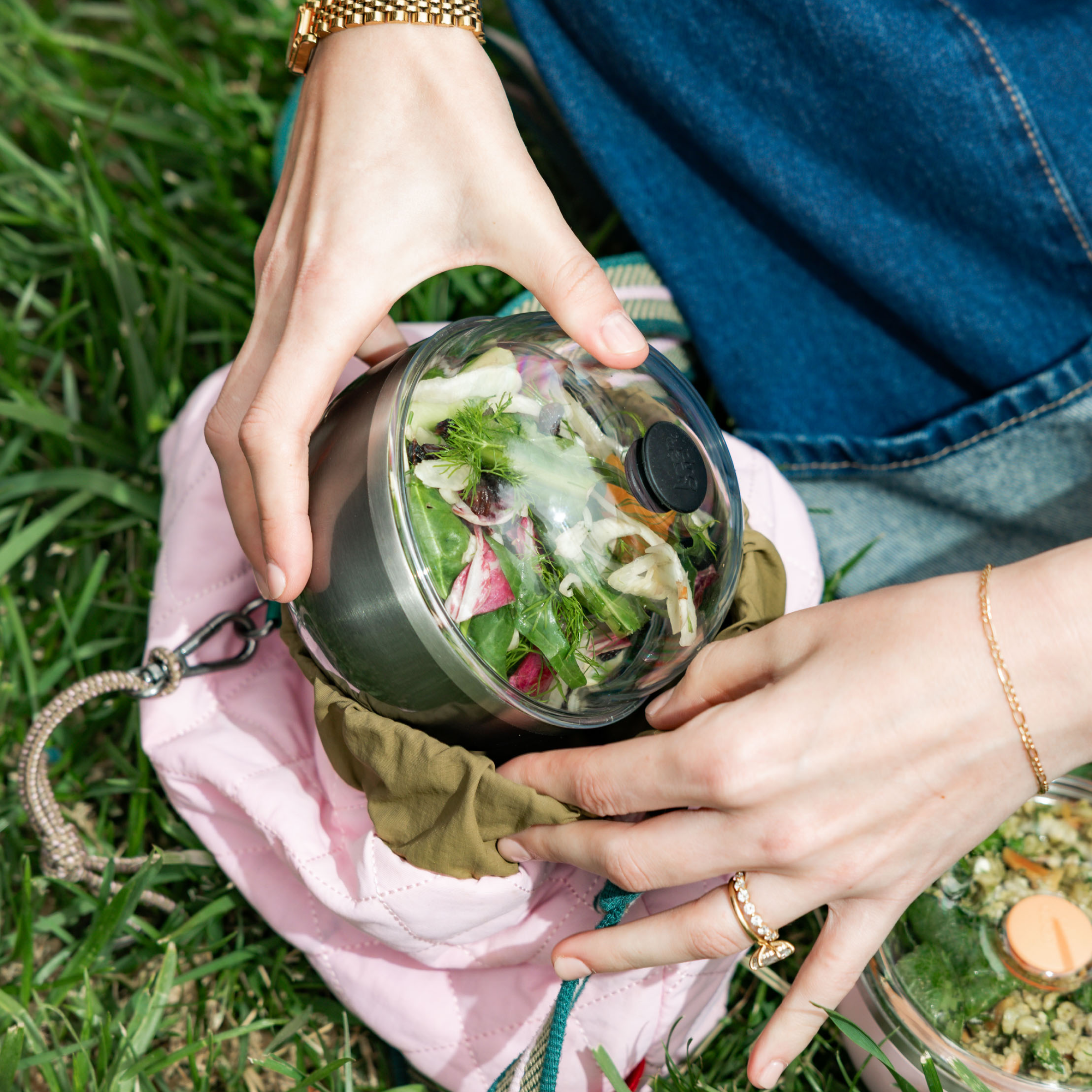 Hands placing a KeepCup Go High Go Low in Nitro colour into a pink quilted bag on grass, the clear domed lid revealing a fresh salad with dill, radish and mixed greens inside.