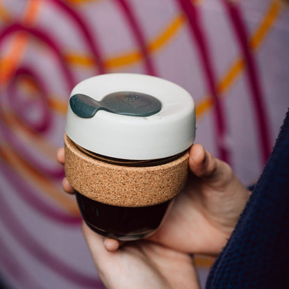 A hand holds a KeepCup Brew Cork S 8oz in Filter, featuring a glass cup with dark coffee visible through the base, natural cork band, and latte lid with soft charocal plug, against a colourful graffiti wall background.