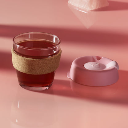 KeepCup Brew Cork XS 6oz in Saskatoon, a glass cup with cork band filled with dark tea or coffee, shown with the peony lid placed beside it on a dusty rose surface with soft lighting and a crystal in the background.