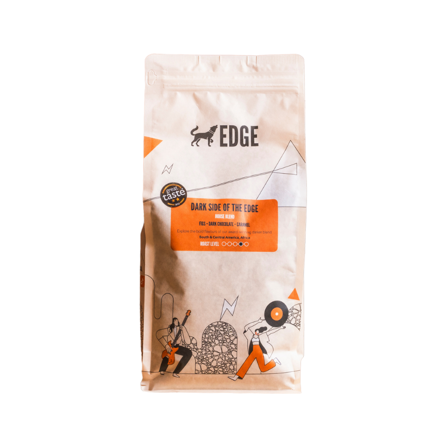 A 1 kg bag of Dark Side of the Edge house blend coffee from Edge, featuring a cream-coloured package with orange accents, illustrated figures playing music, and tasting notes of figs, dark chocolate and caramel displayed on the front label.