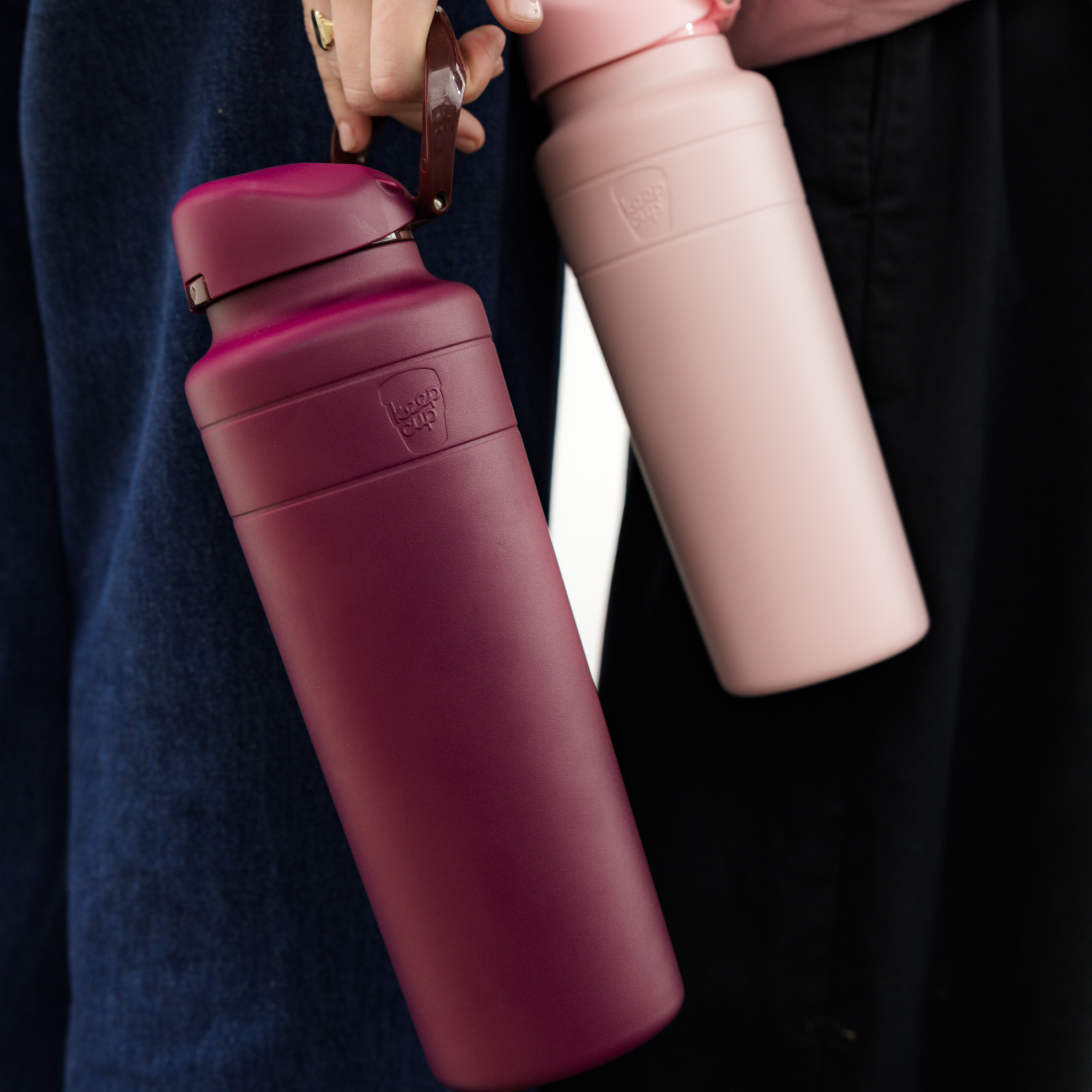 Two insulated water bottles, one pink and one mulberry, held by a person against a dark background.