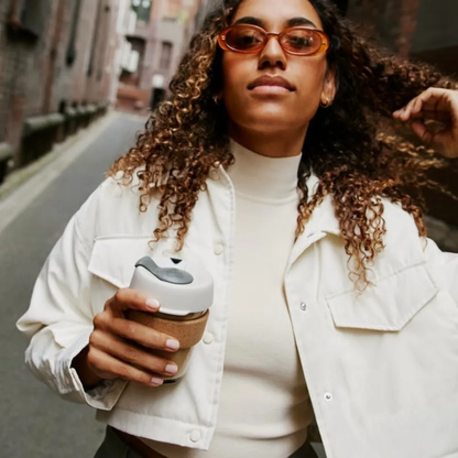 A woman with curly hair and orange sunglasses walks down an urban street holding a KeepCup Brew Cork M 12oz in Filter colourway, featuring a soft charcoal lid and natural cork band, paired with a cream turtleneck and white jacket.